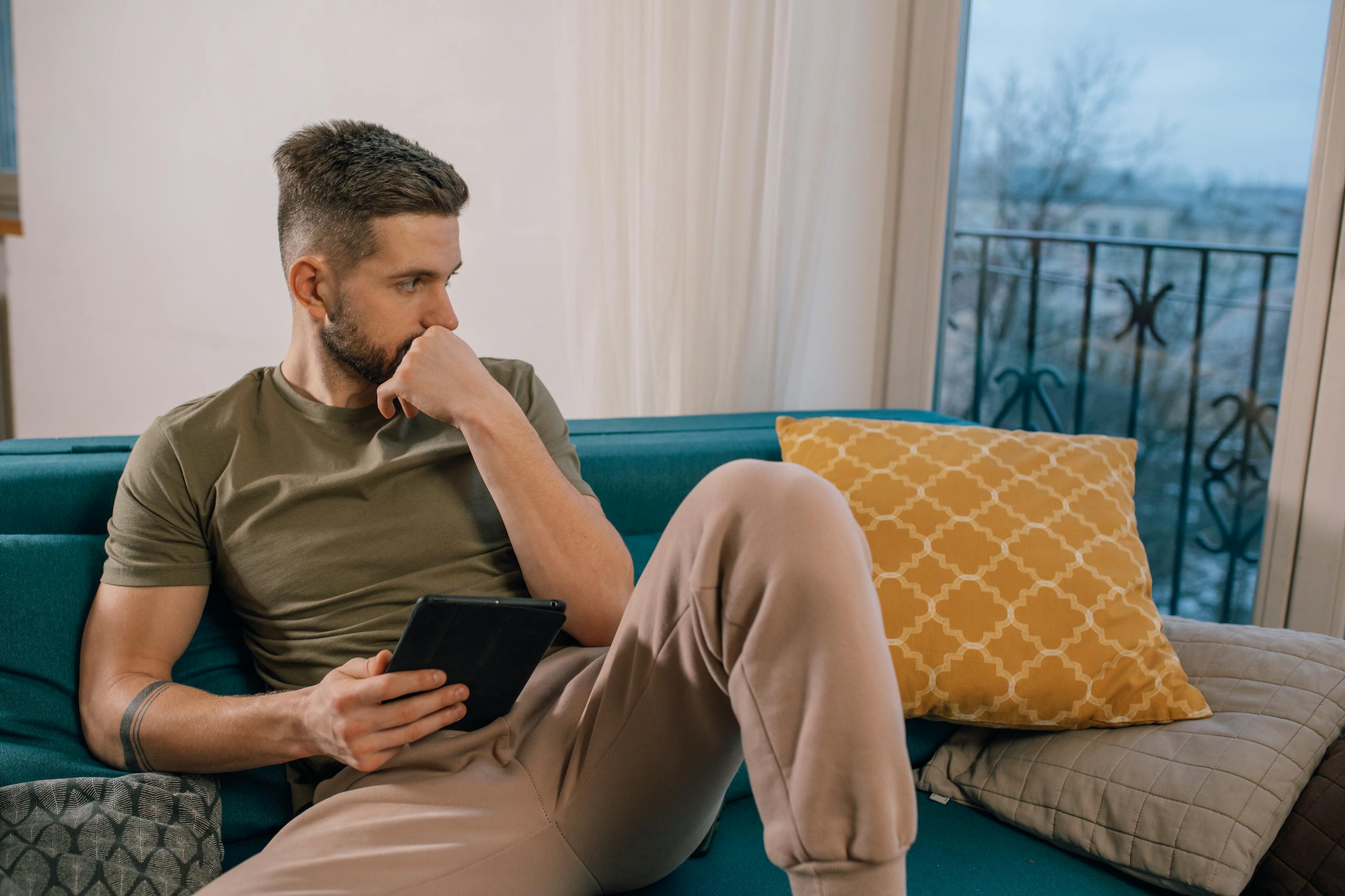 Bearded man sitting on a sofa, thoughtfully holding a tablet indoors.